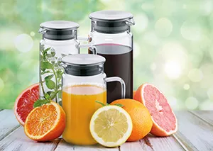 Glaskaraffen Set für Saft und Getränke mit frischen Früchten Glaskaraffen mit Säften, frischen Zitrusfrüchten und Kräutern auf Holztisch vor unscharfem Hintergrund
