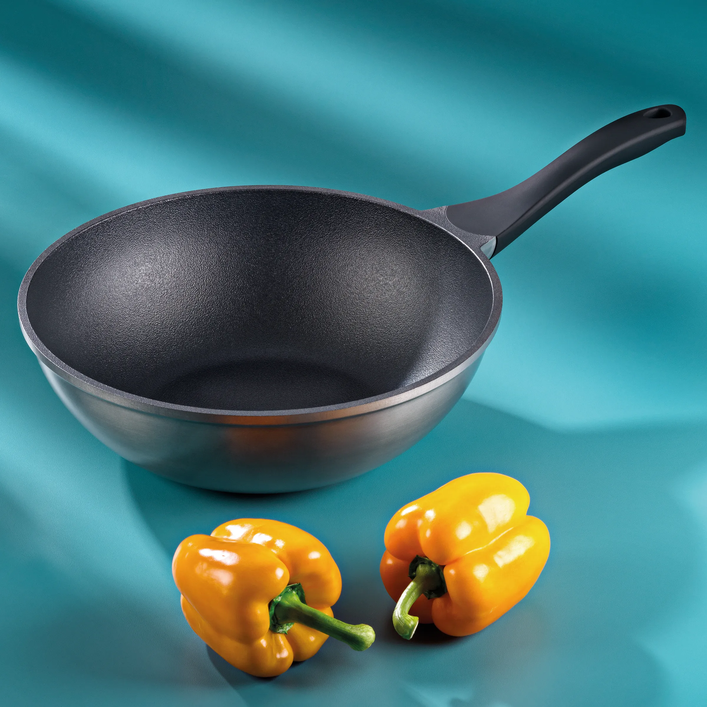 Antihaft-Bratpfanne mit schwarzer Beschichtung und grauem Griff, daneben zwei gelbe Paprika auf türkisem Hintergrund