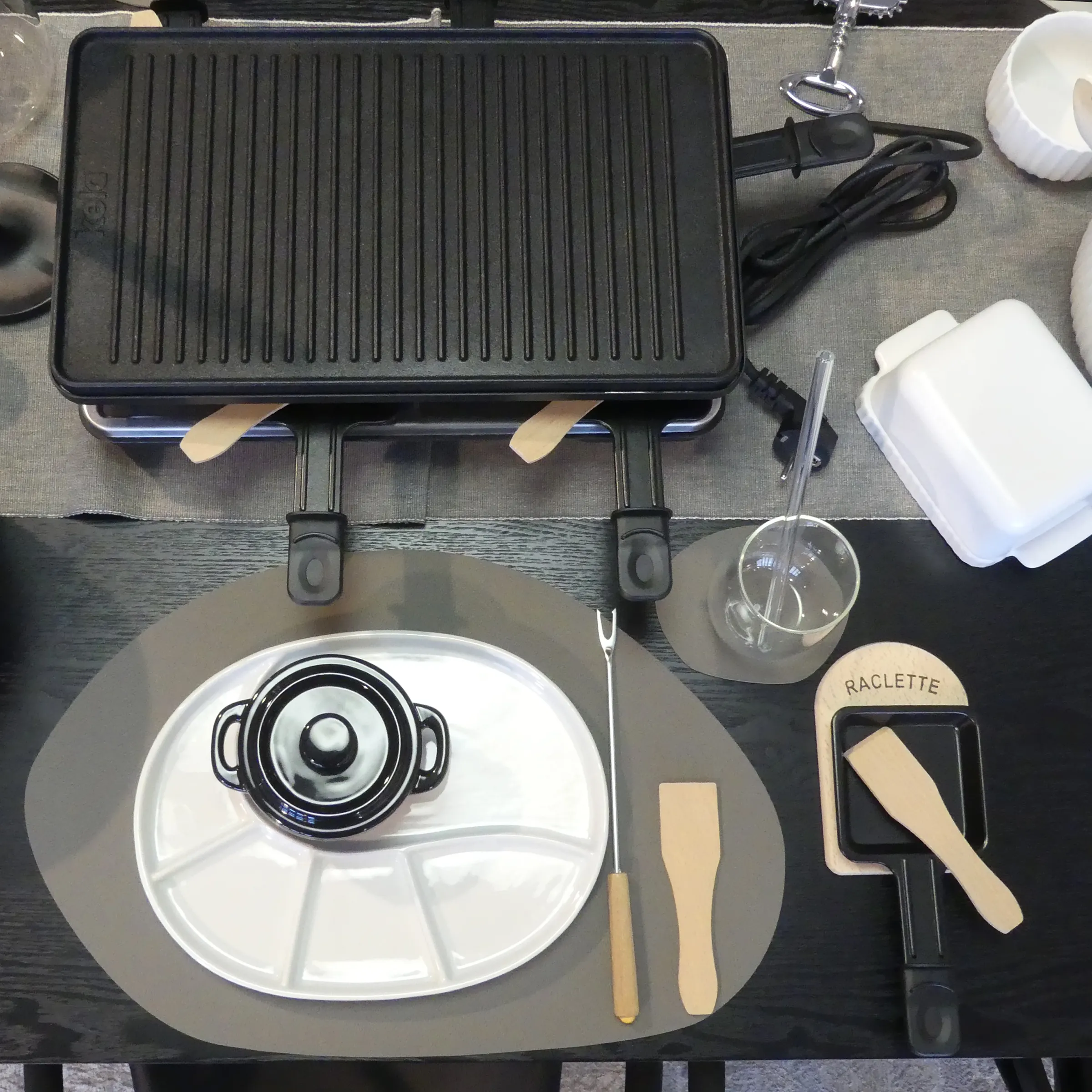 Raclette-Grill Set mit Pfännchen und Zubehör - Komplette Tischausstattung Raclette-Grill mit Zubehör auf gedecktem Tisch mit Pfännchen, Tellern und Holzspateln