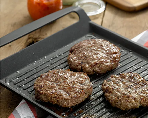 Drei Fleischbulletten braten in einer schwarzen Grillpfanne auf Holzuntergrund neben Gewürzen und Saucen