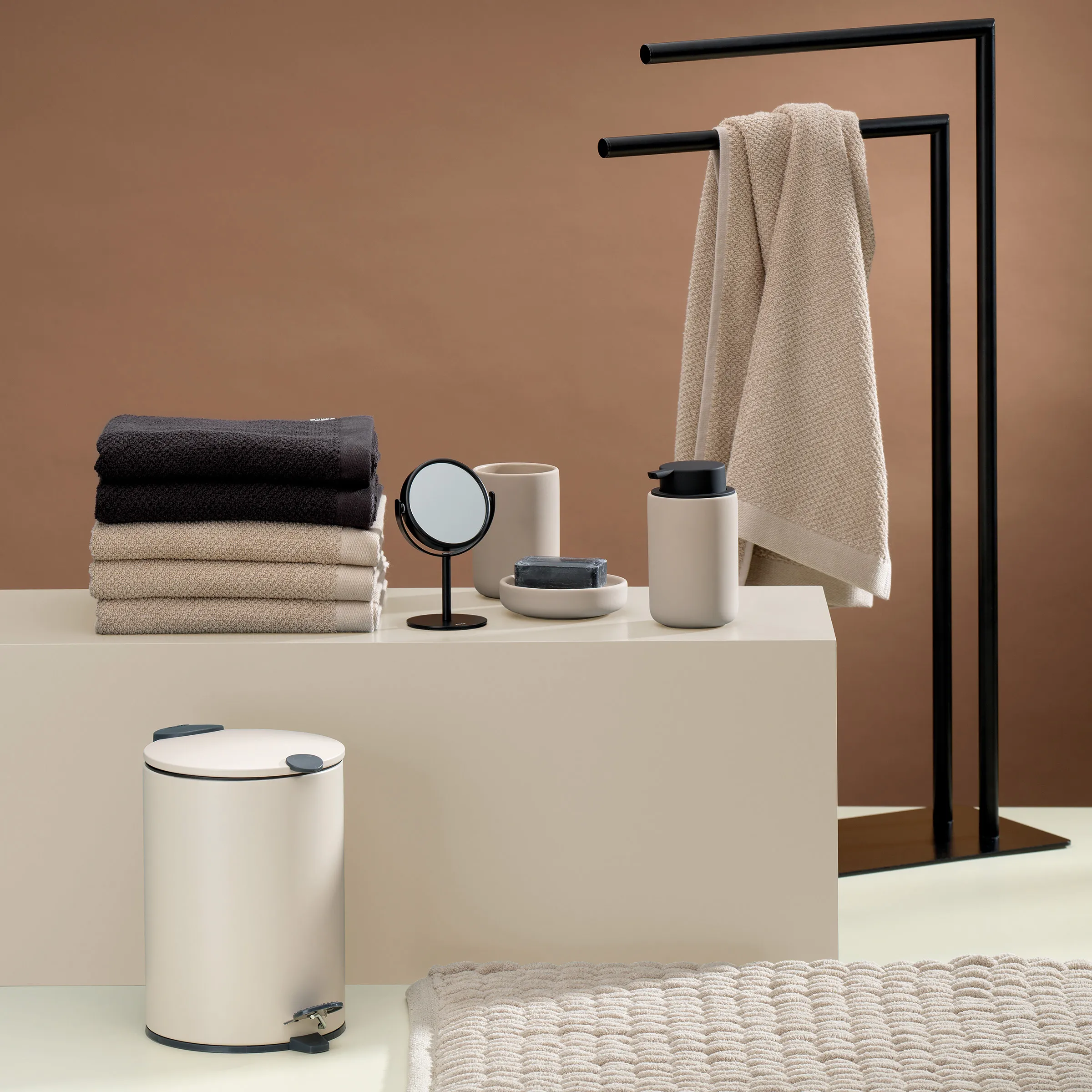 Modernes Badezimmer-Set mit gestapelten Handtüchern, Kosmetikeimer, Seifenspender, Spiegel und Handtuchhalter mit Tuch vor braunem Hintergrund.