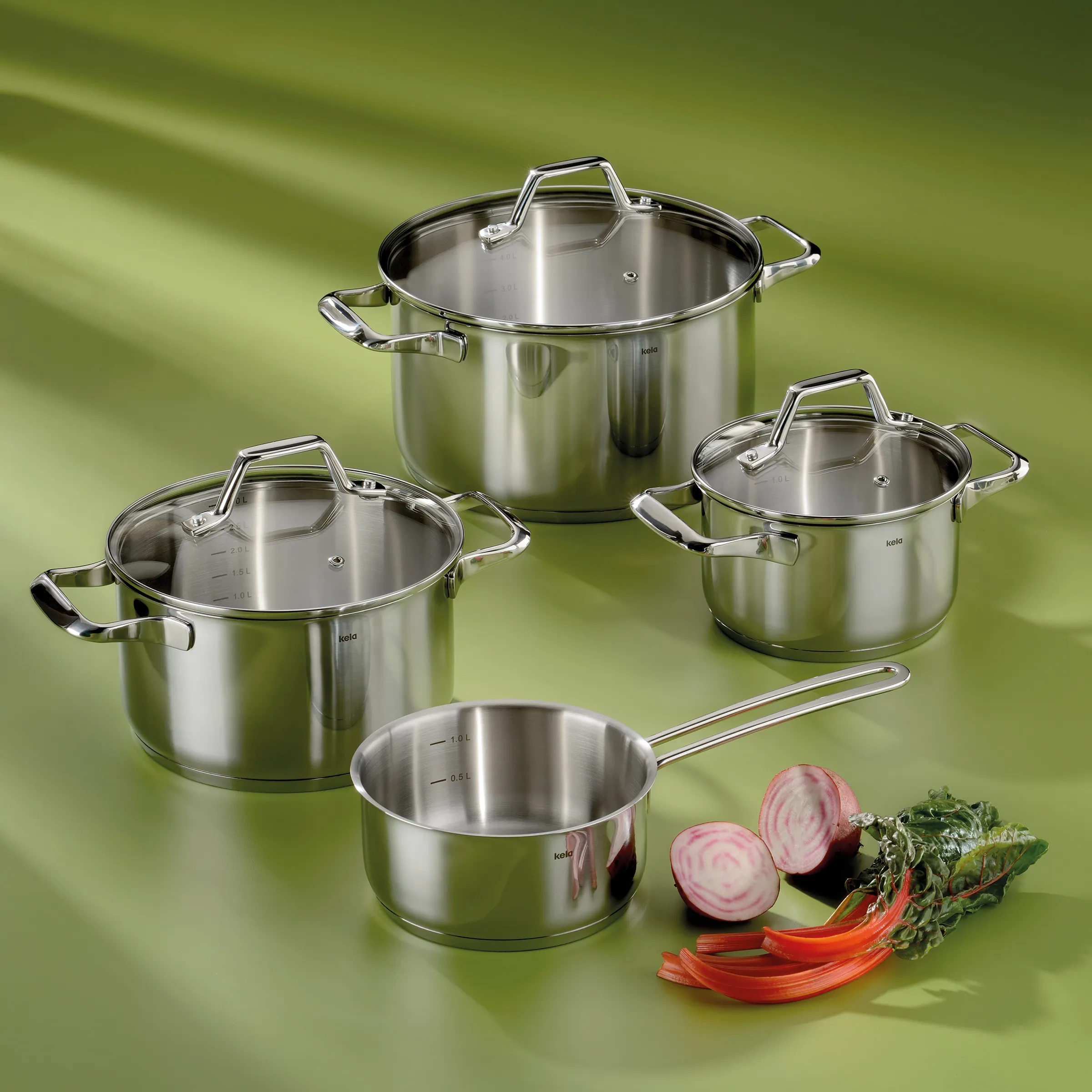 Stainless steel cookware set featuring three pots in various sizes with glass lids and a saucepan, displayed with fresh vegetables.