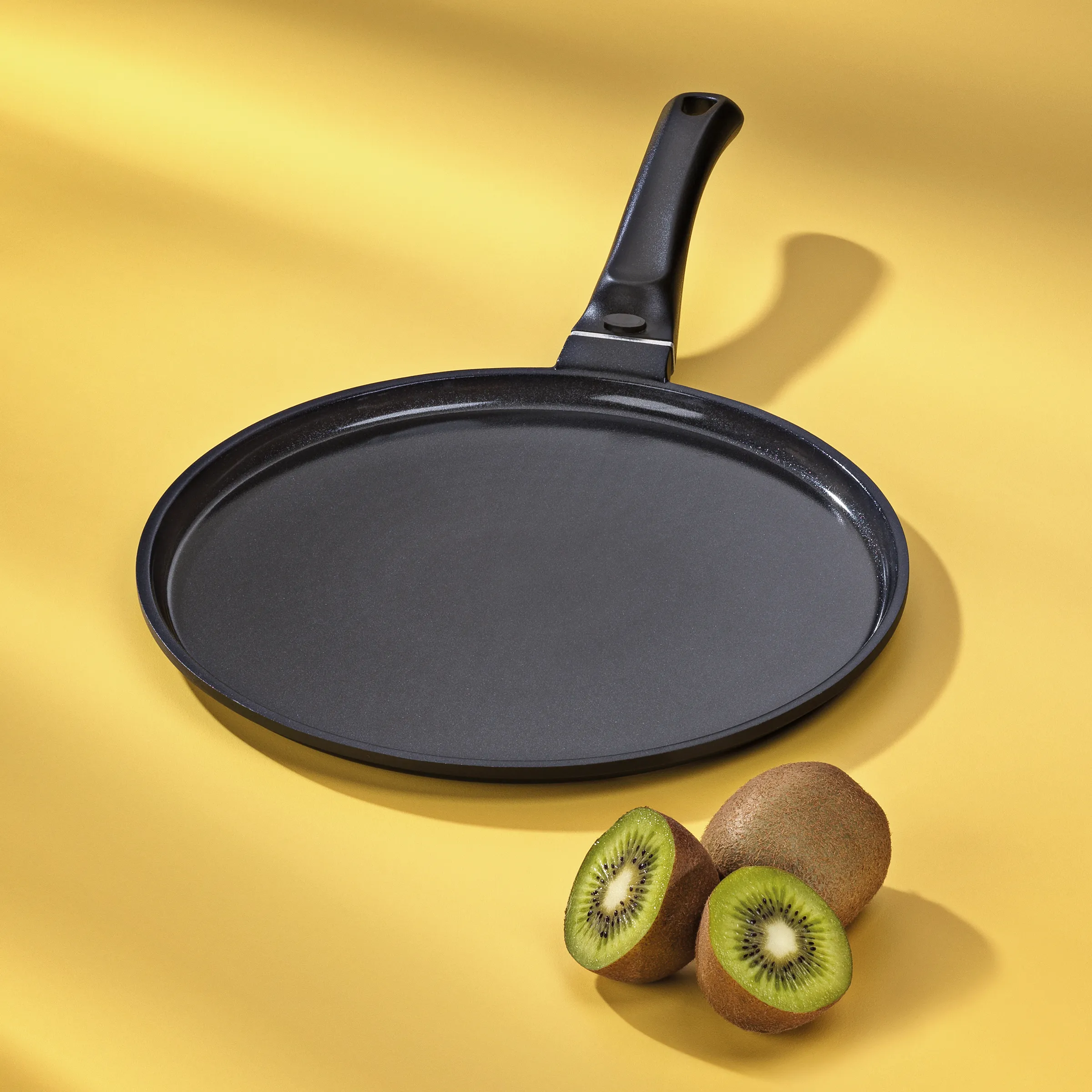 Crepes-Pfanne antihaftbeschichtet - Flache Bratpfanne Schwarze Crepes-Pfanne mit schwarzem Griff auf gelbem Hintergrund, daneben aufgeschnittene Kiwis