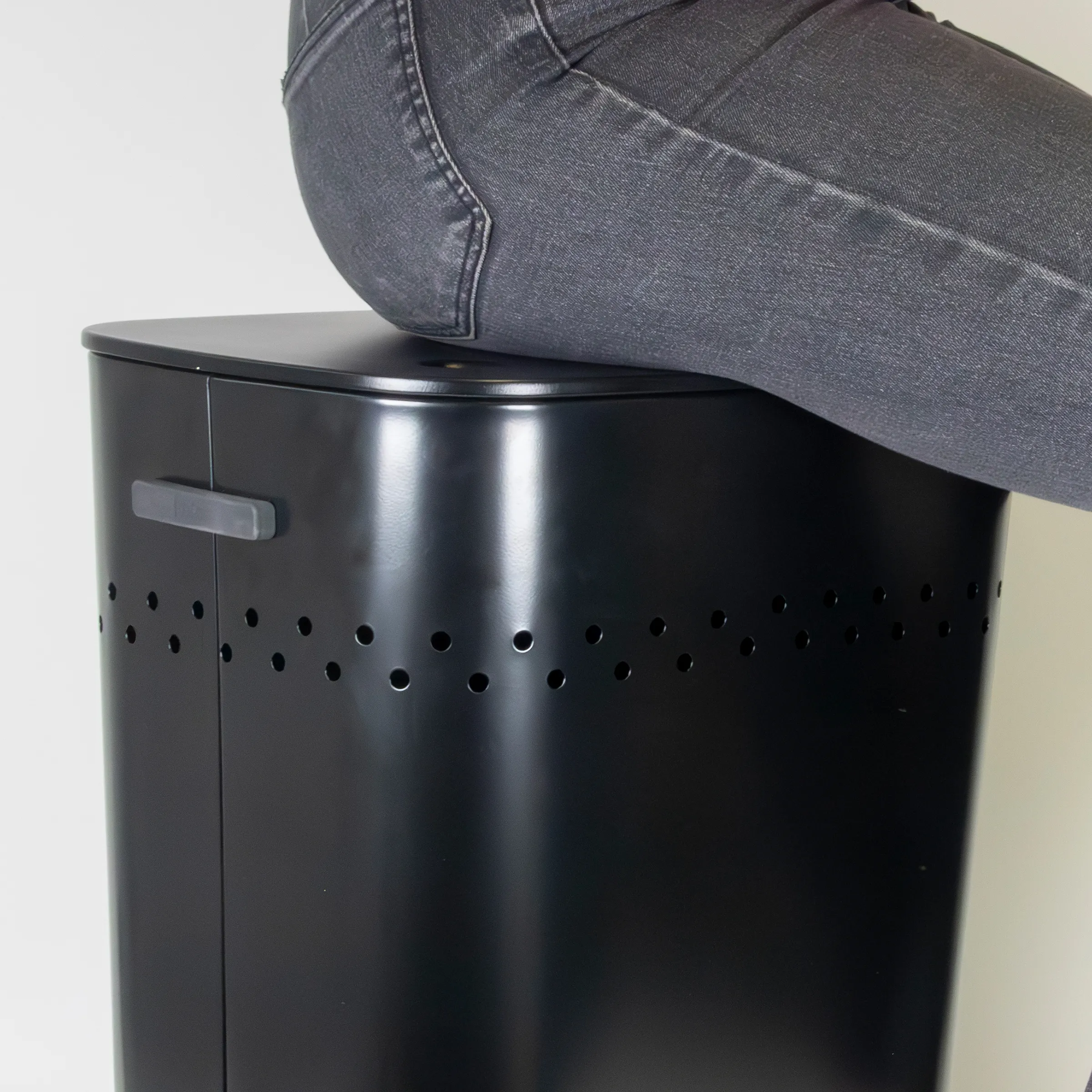 Person sitzt auf schwarzem Metallhocker mit Belüftungslöchern und grauer Denim-Hose