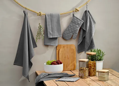 Graue Küchenhandschuhe, Handtücher und Topflappen hängen an Seil. Darunter Holzbrett, Pflanzen und Deko-Accessoires auf Holztisch.