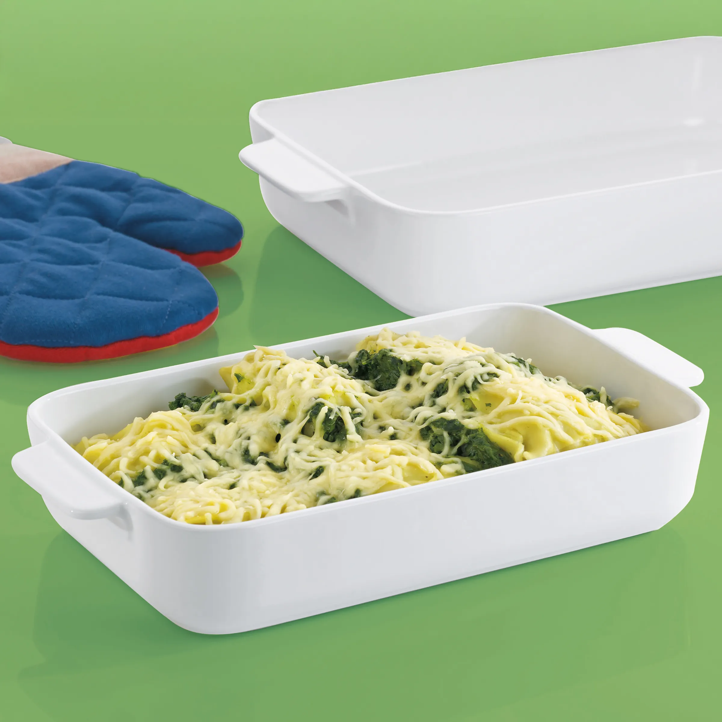 Auflaufform Pasta Gratin mit Spinat Weiße Auflaufform mit Pasta und grünem Blattgemüse auf grünem Hintergrund neben blauen Ofenhandschuhen