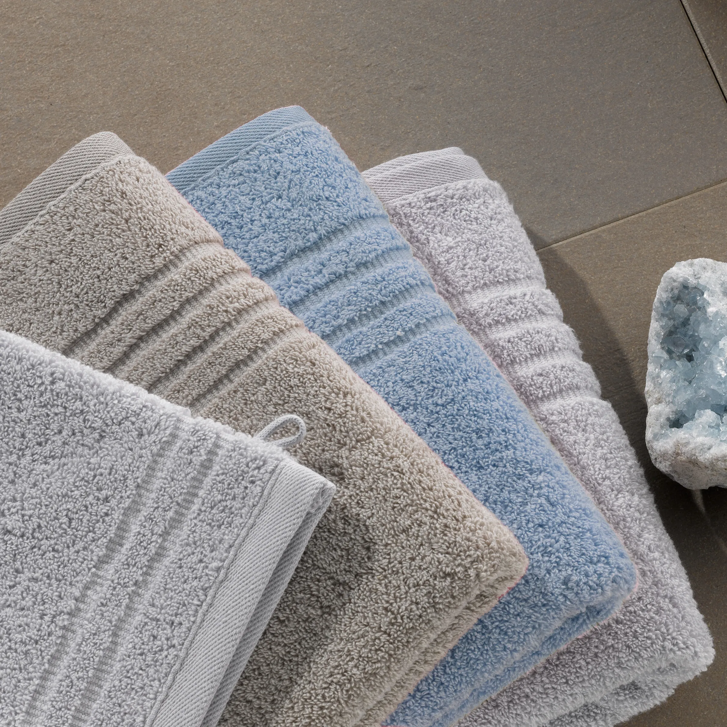 Vier gefaltete Handtücher in verschiedenen Farben - Weiß, Beige, Blau und Grau - gestapelt auf beigem Untergrund