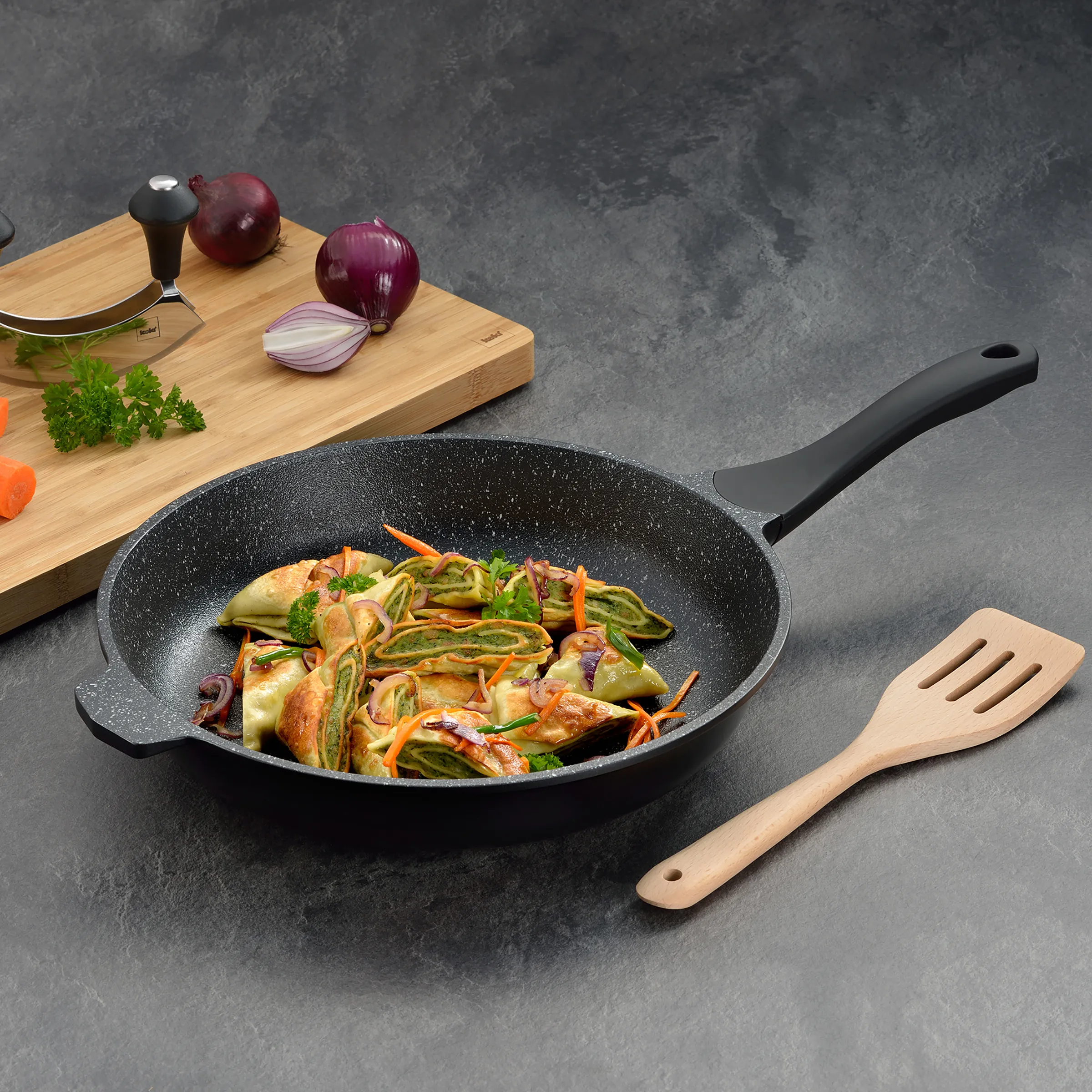 Wok-Pfanne mit Gemüse - Antihafte Bratpfanne für Wokgerichte Schwarze Wok-Pfanne mit Gemüse und Fleisch auf grauem Hintergrund, daneben Holzschneidebrett mit Zwiebeln und Kräutern