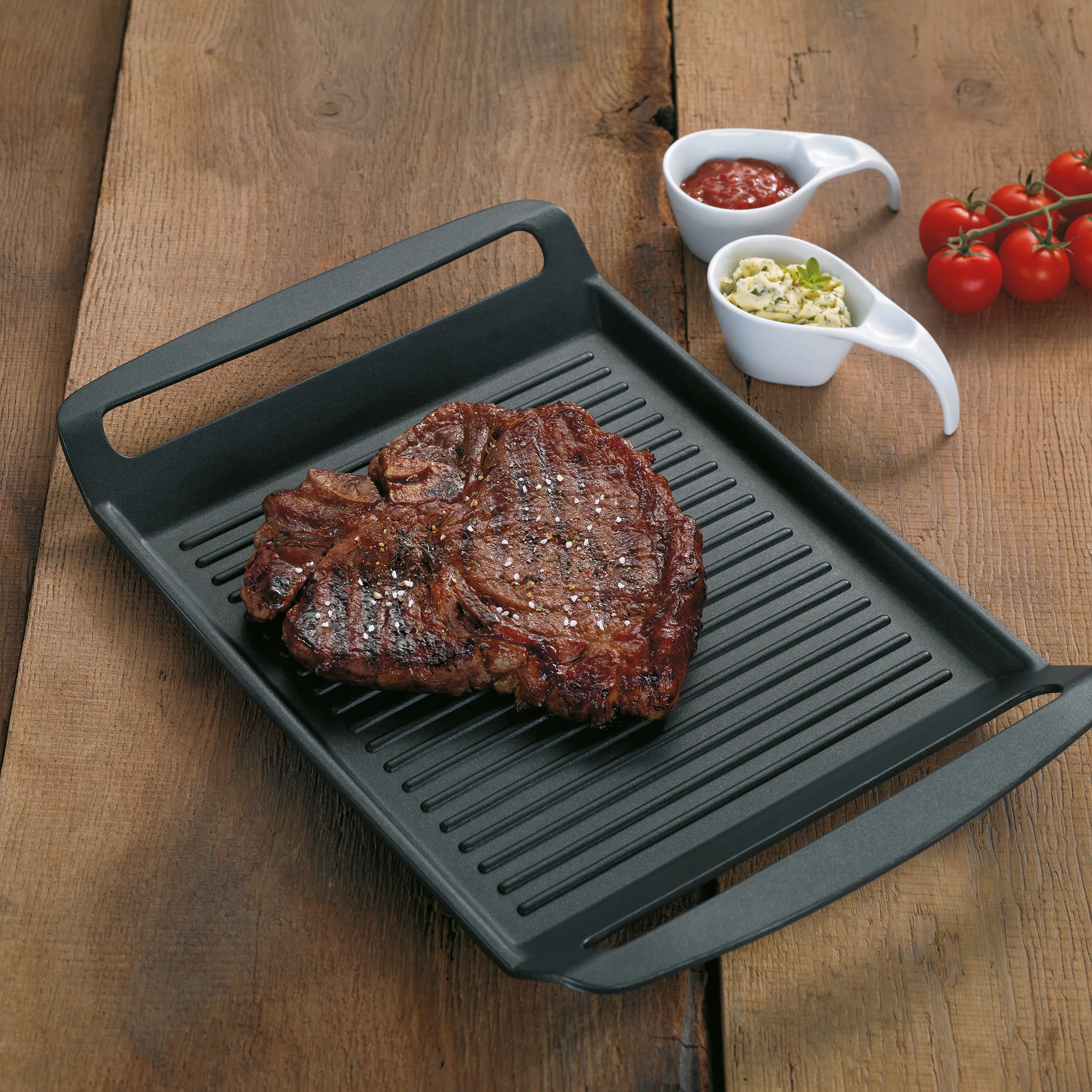 Grillpfanne mit Steak und Gewürzen - Grill-Zubehör für perfektes Fleisch Gegrilltes Steak auf schwarzer Grillpfanne mit Ketchup, Kräuterbutter und Tomaten auf Holztisch