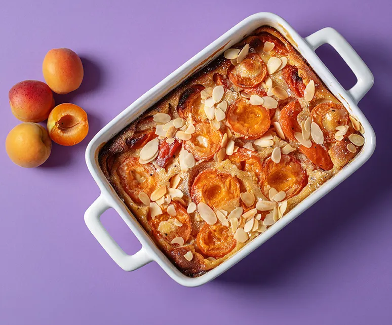 Aprikosen-Mandelkuchen Rezept mit frischen Früchten Aprikosen-Mandelkuchen in weißer Backform mit frischen Aprikosen auf violettem Hintergrund