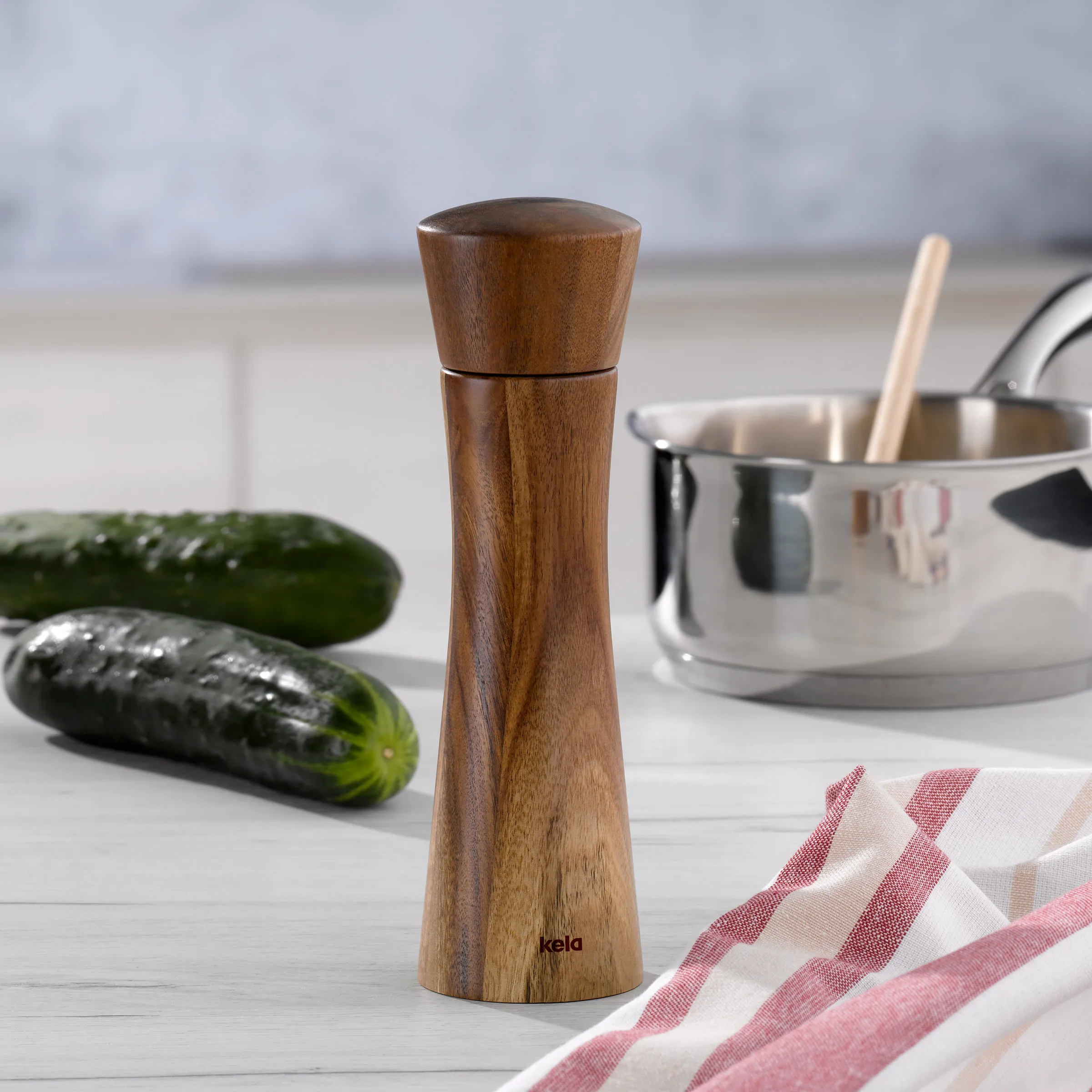 Holzpfeffermühle aus dunklem Holz auf weißer Küchenarbeitsfläche mit Gurken und Kochtopf im Hintergrund