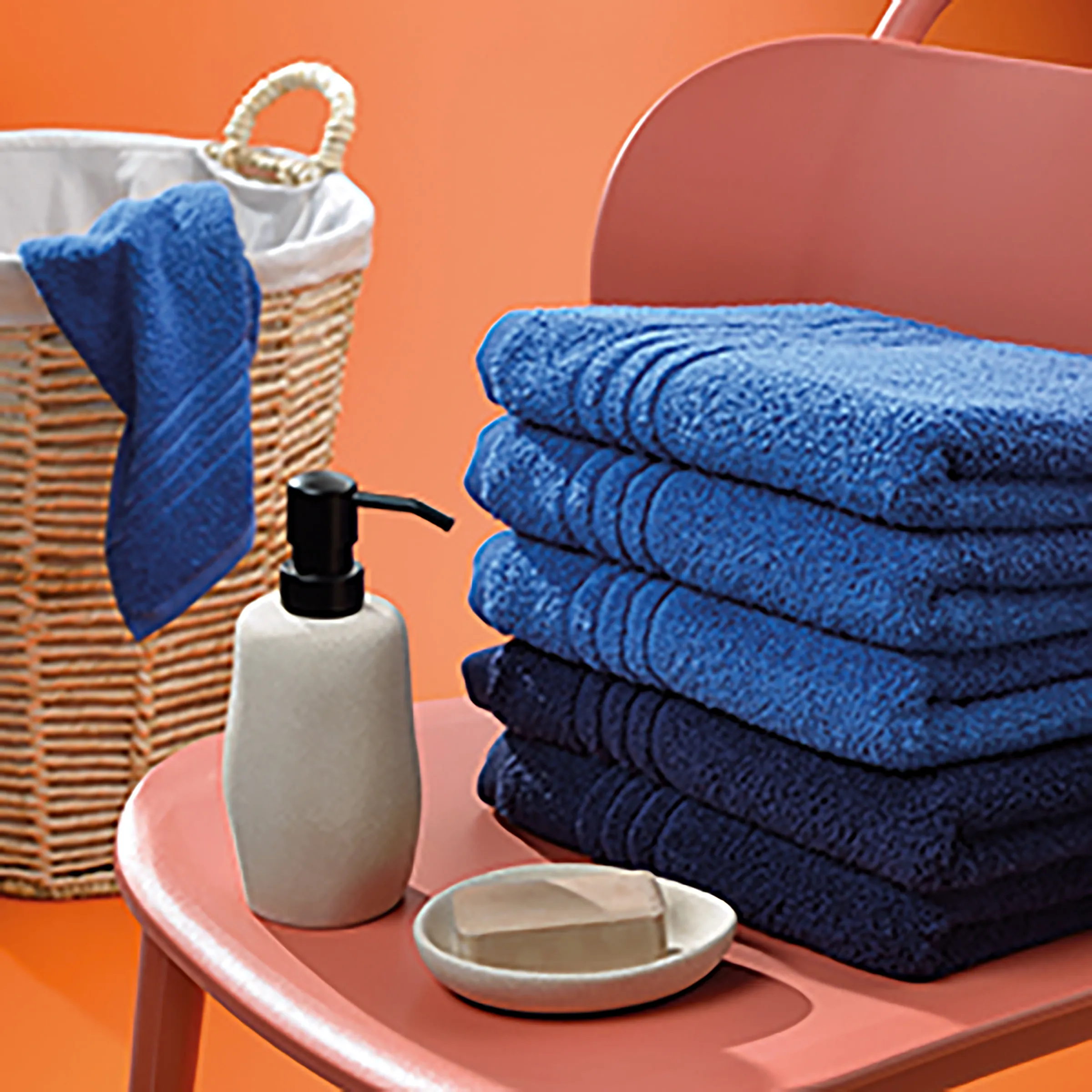 Badezimmer Handtücher Set mit Accessoires Badezimmer-Arrangement mit gestapelten blauen Handtüchern, Seifenspender, Seifenschale, Weidenkorb und rotem Stuhl auf orangefarbenem Hintergrund