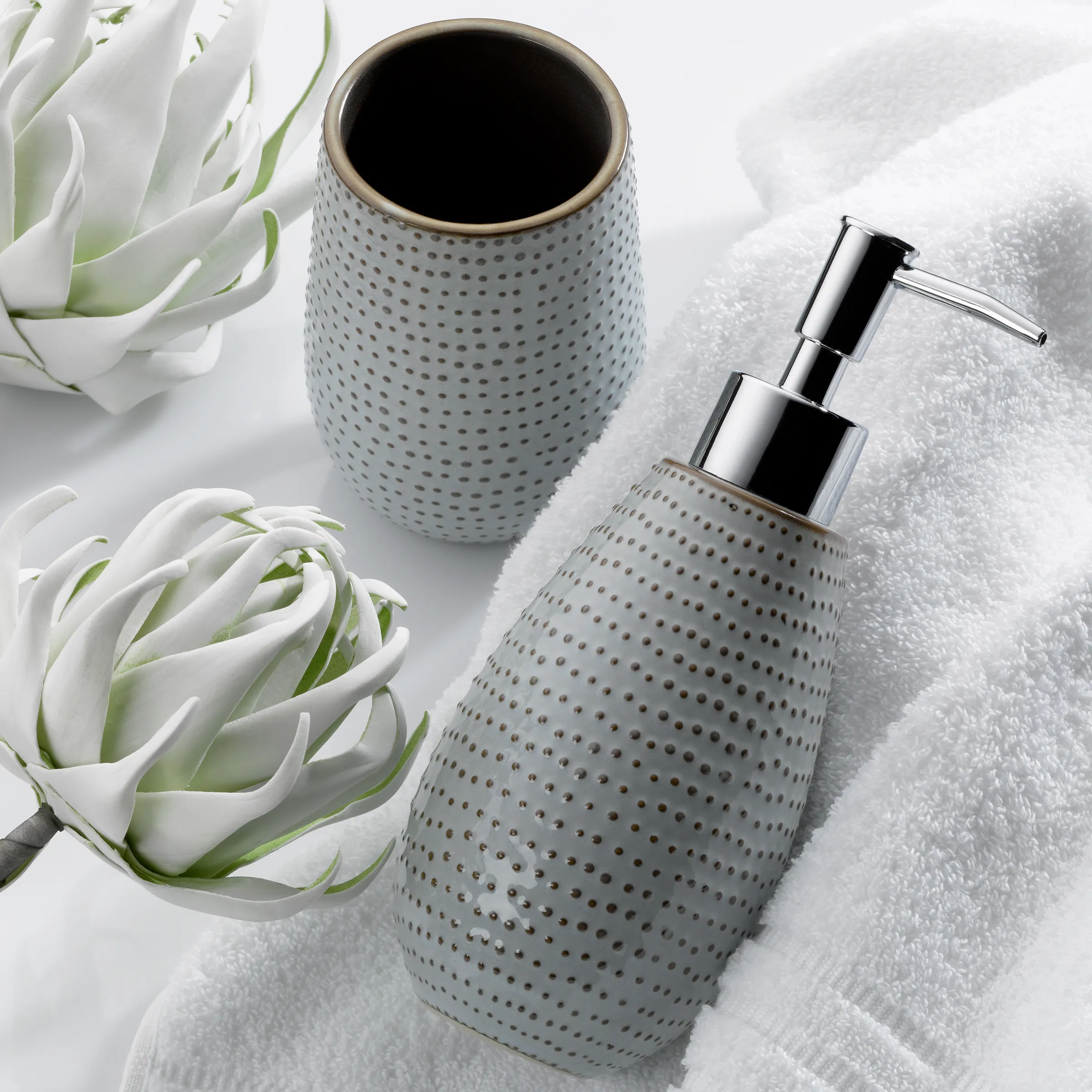 Moderne Badaccessoires Set Keramik mit Seifenspender Graue Keramik-Badaccessoires mit Punktmuster: Seifenspender und Zahnbürstenhalter neben weißen Sukkulenten auf Handtuch