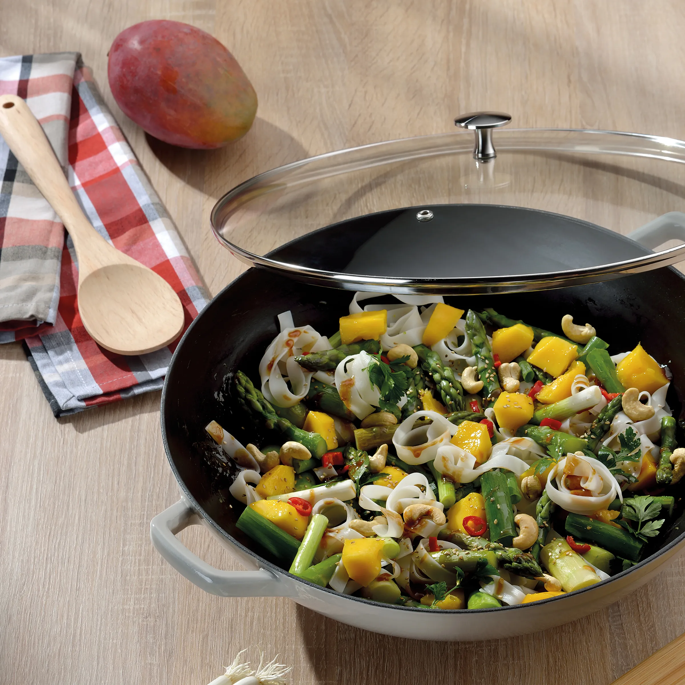 Wok Pfanne mit Gemüse Nudeln Rezept Wok mit Gemüse-Nudelgericht, Holzlöffel, rot-weiße Serviette und Mango auf Holztisch