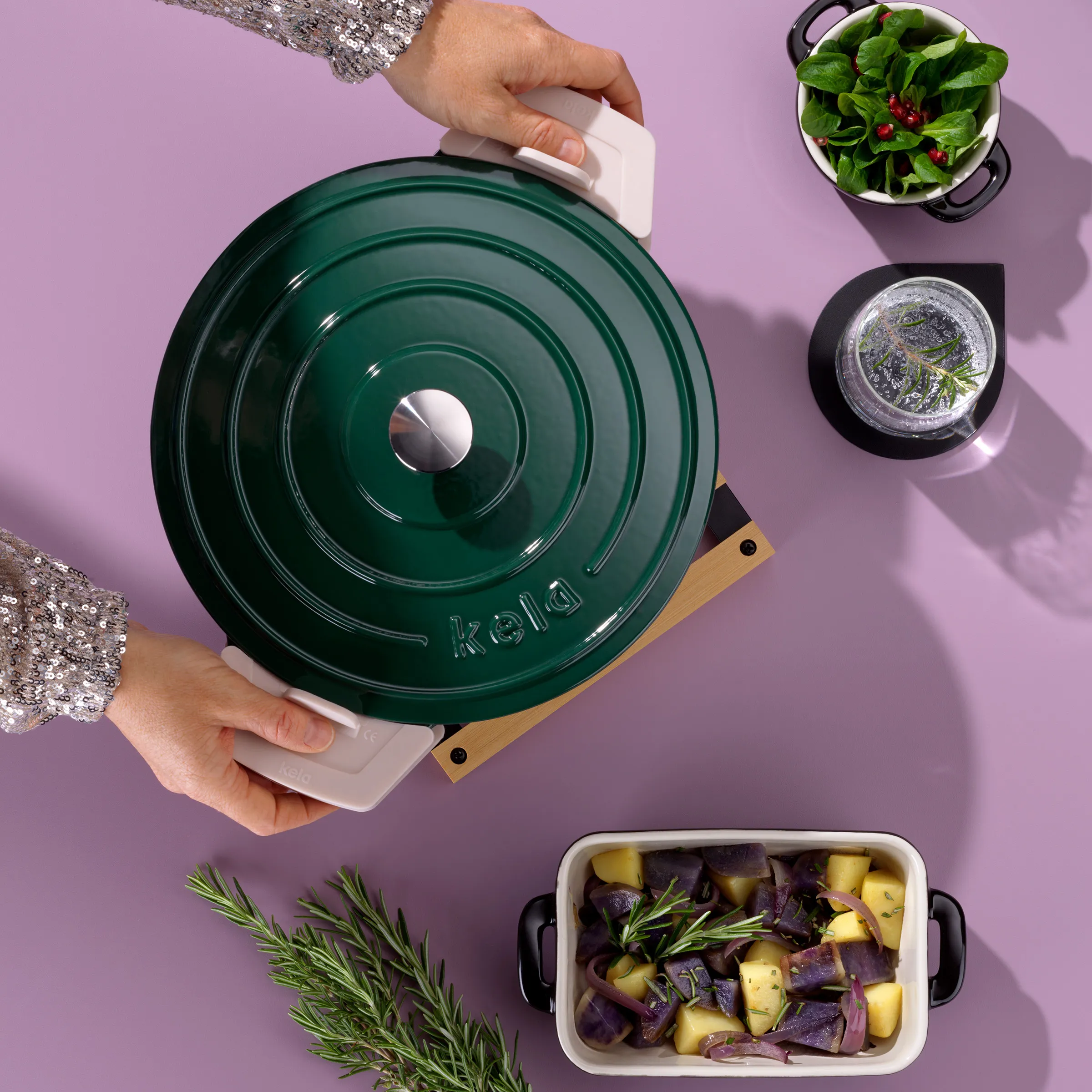 Kela Gusseisentopf mit Deckel - Premium Kochtopf für gesunde Küche Hände halten grünen Kela Gusseisentopf mit Deckel über rosa Hintergrund, umgeben von frischen Kräutern und Gemüse
