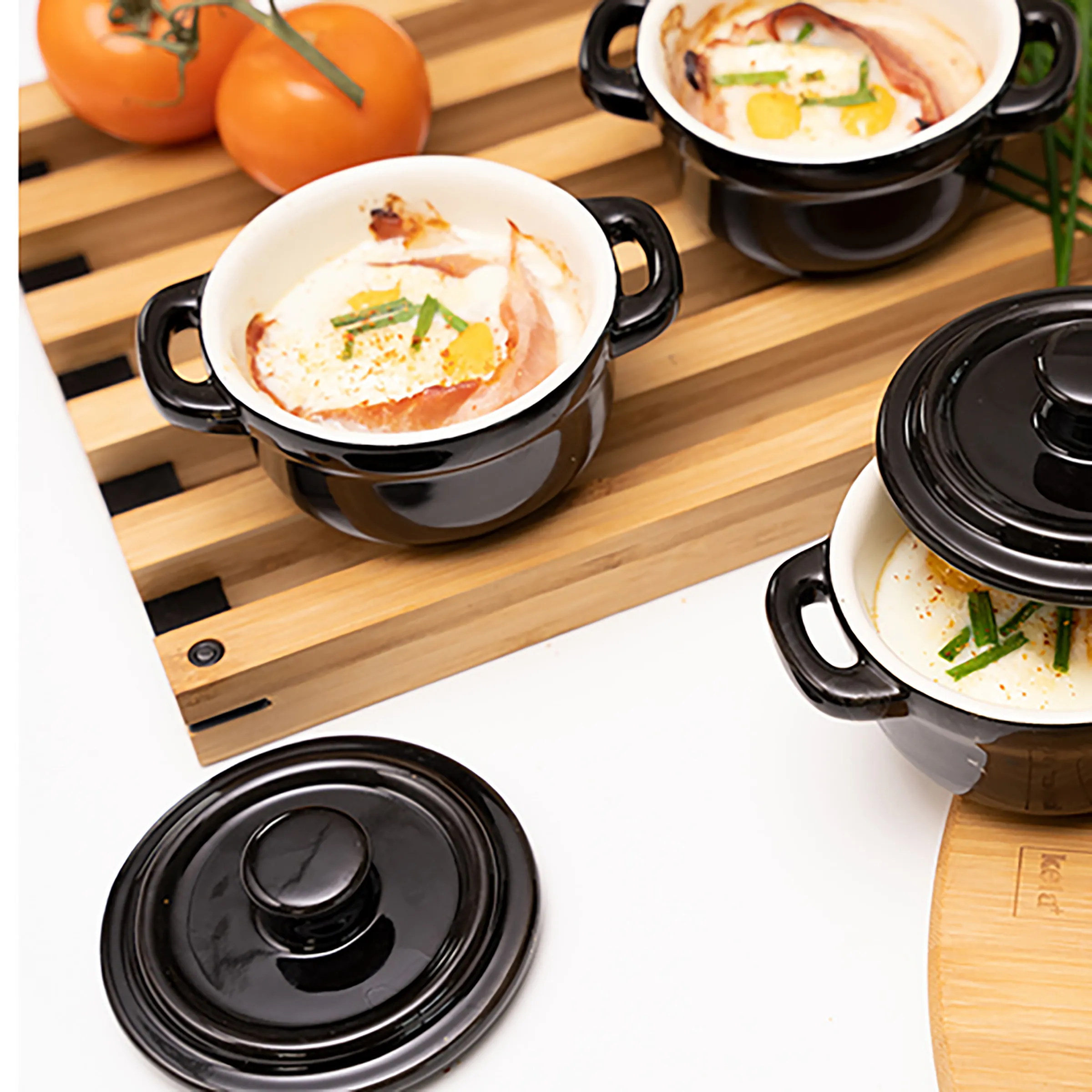 Elegante Keramiktöpfe für Suppen und Eintöpfe Schwarze Keramiktöpfe mit Griffen und Deckeln auf Holzbrett, gefüllt mit Cremesuppe, Lachs, Gemüse und Kräutern garniert, frische Tomaten im Hintergrund.
