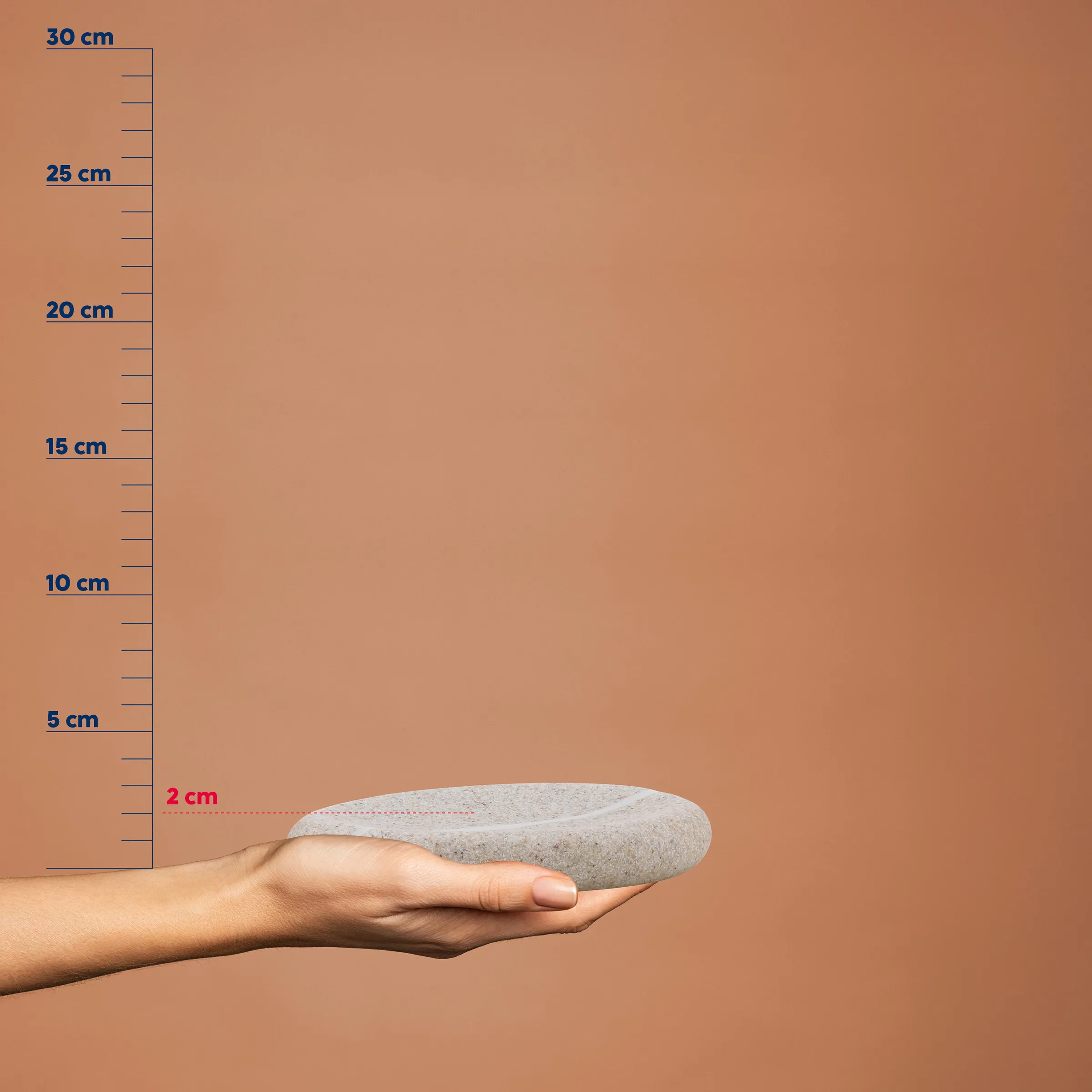 Hand hält einen grauen Stein mit Größenvergleichsskala von 2-30 cm an braunem Hintergrund