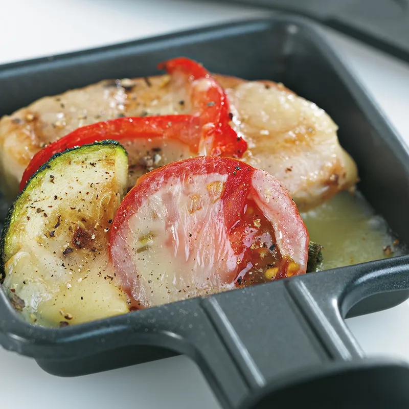 Raclette-Pfännchen mit Gemüse und Käse – Raclette-Zubereitung Raclette-Pfännchen mit geschmolzenem Käse, Zucchini, Paprika und Gewürzen in schwarzer Raclette-Schale