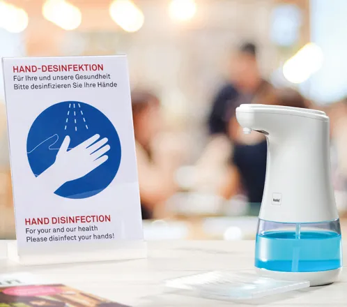 Automatischer Desinfektionsspender neben Hinweisschild mit Anleitung zur Handdesinfektion in Büro