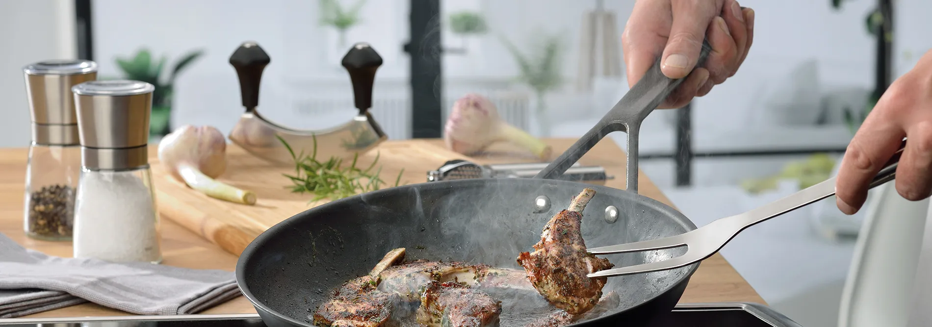 Lammkoteletts in Granitsteinpfanne braten - Küchenzange und Kochgeschirr Hand hält Grillzange mit Lammkotelett über Granitsteinpfanne beim Braten in moderner Küche