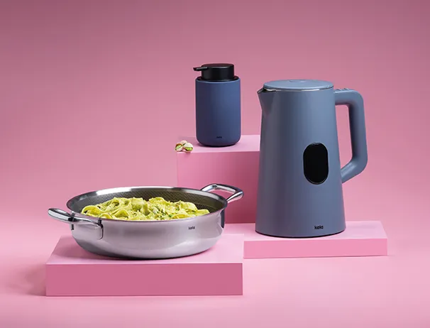 Küchenset mit blauem Wasserkocher, blauer Thermoskanne und Edelstahlpfanne mit Nudeln auf rosa Podesten