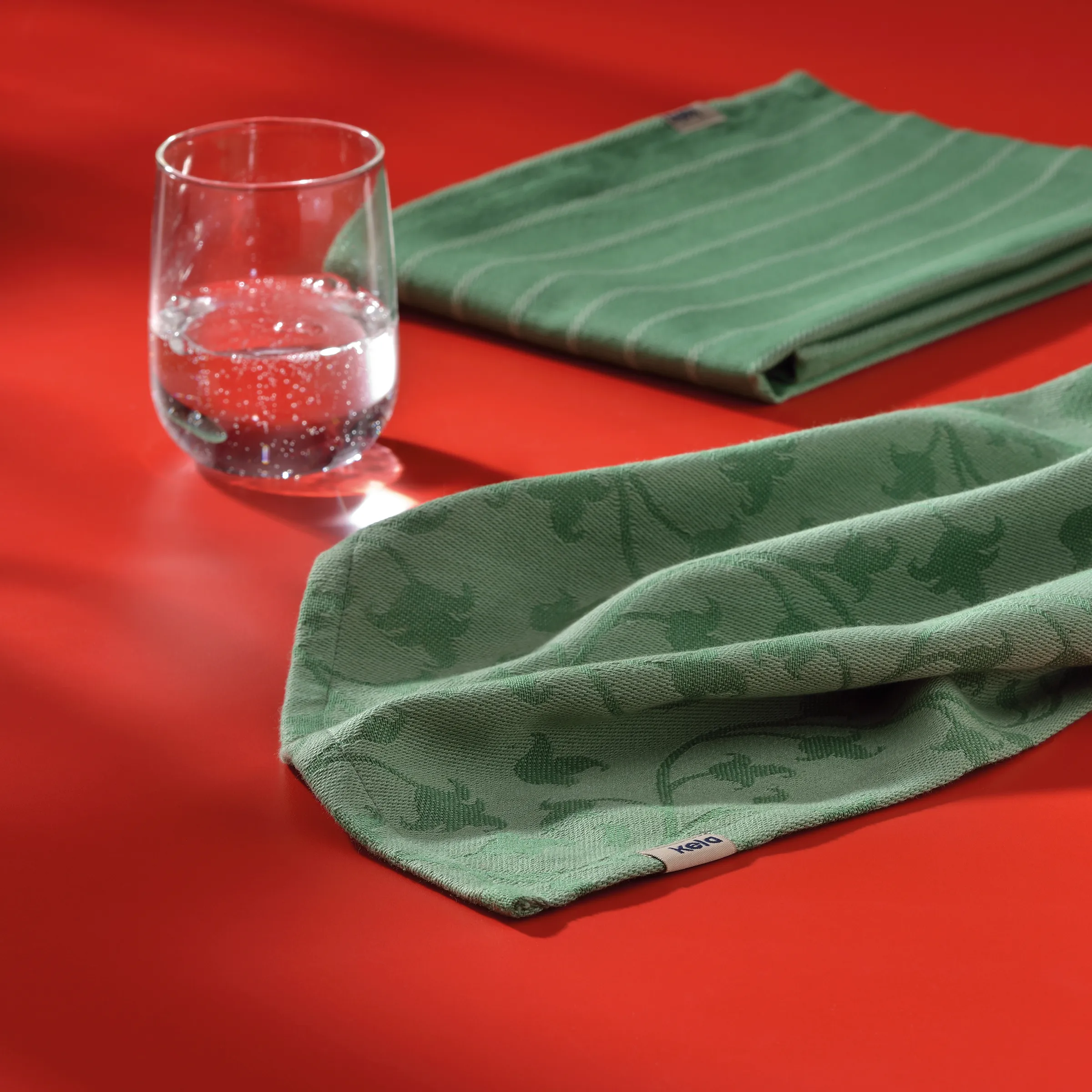 Glas Wasser mit grünem Handtuch - Erfrischung Design Klares Glas mit Wasser und Blasen auf rotem Hintergrund neben grünen Handtüchern mit Muster