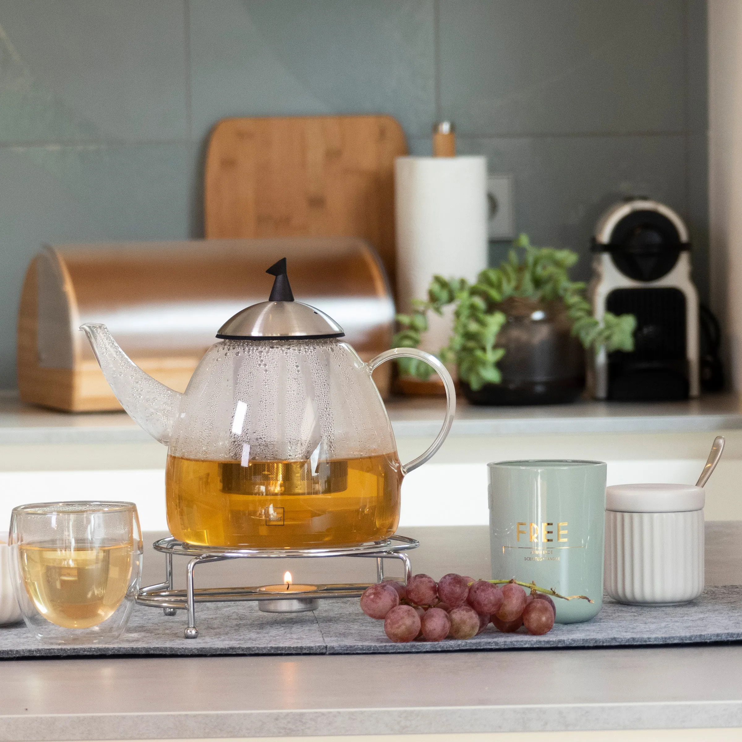 Teekanne mit goldenem Tee auf Stövchen, daneben Teeglas, Teedose und Weintrauben auf Schieferplatte