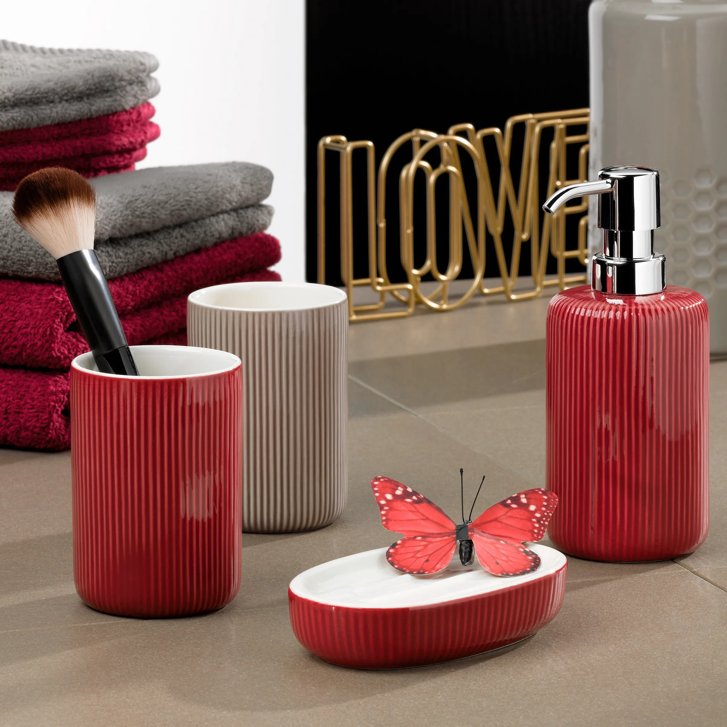 Badezimmer-Accessoires Set Rot - Moderne Bad-Ausstattung mit Seifenspender Rotes Badezimmer-Accessoire-Set mit Seifenspender, Becher und Seifenschale auf grauer Oberfläche mit Handtüchern