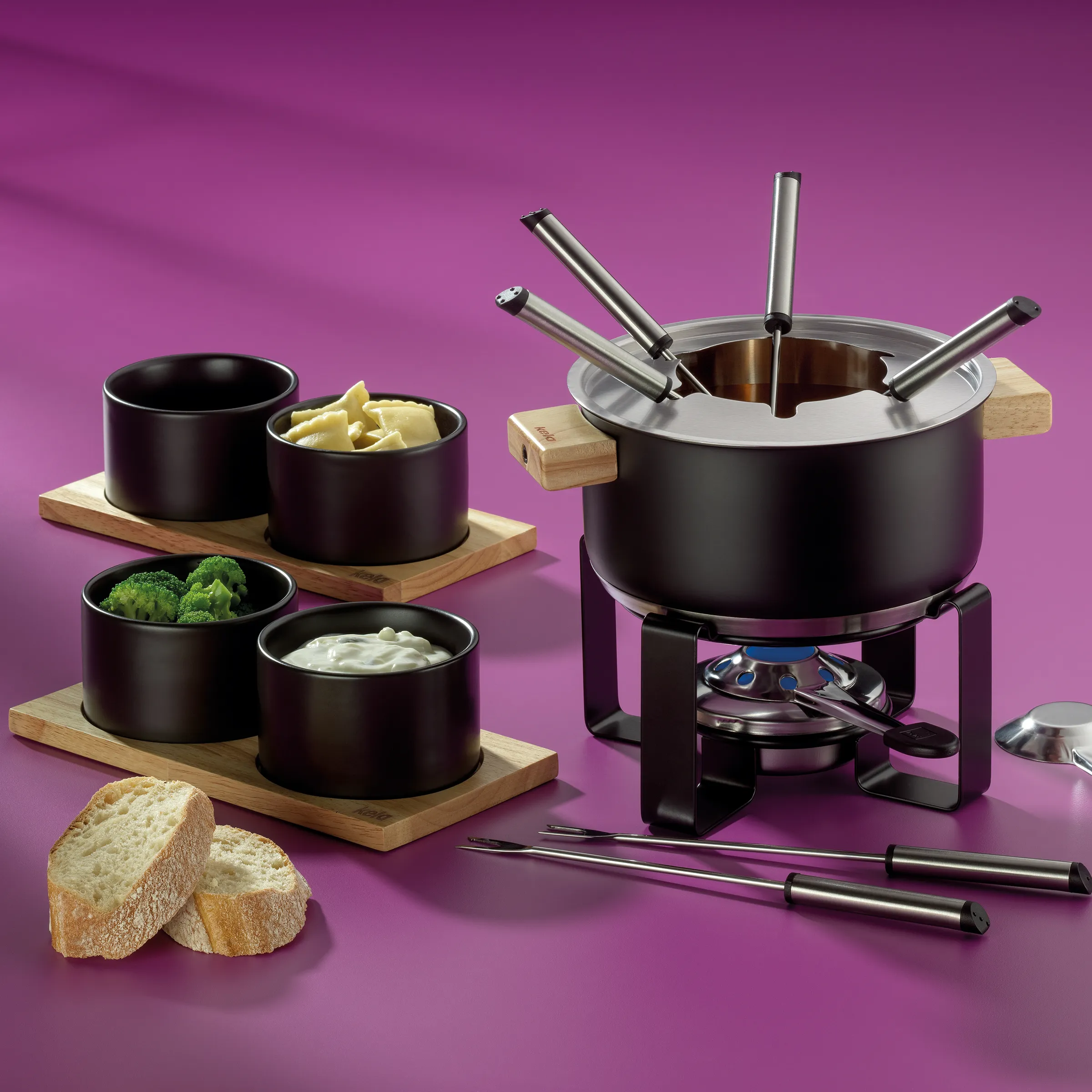 Käse-Fondue-Set: Komplettes Fondue-Service mit Topf und Zubehör Käse-Fondue-Set mit schwarzem Topf, vier Porzellanschüsseln mit Zutaten und Holzbrettern auf rosa Hintergrund