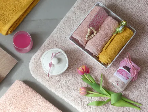 Spa-Set mit Handtüchern in Pastellfarben, Blüten, Kosmetikprodukten und dekorativen Elementen auf grauer Oberfläche.