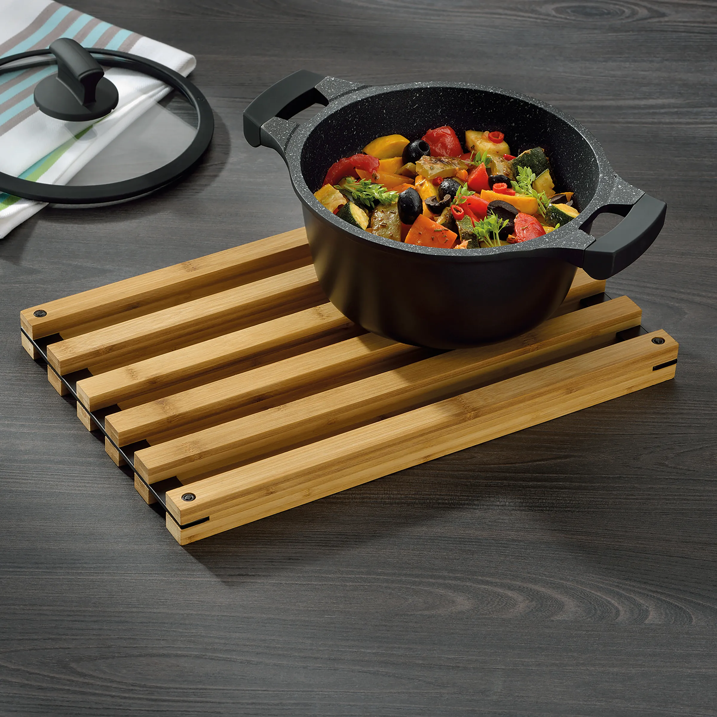 Kochtopf mit Deckel und Bamboo-Untersetzer - Kochgeschirr Set Schwarzer Kochtopf mit Gemüse auf Bamboo-Untersetzer, Topfdeckel und Serviette im Hintergrund