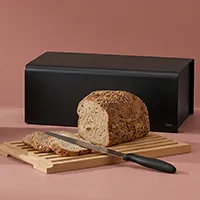 Brotkasten aus Metall in Schwarz mit geschnittenem Vollkornbrot, Messer und Holzbrett auf rosafarbenem Hintergrund