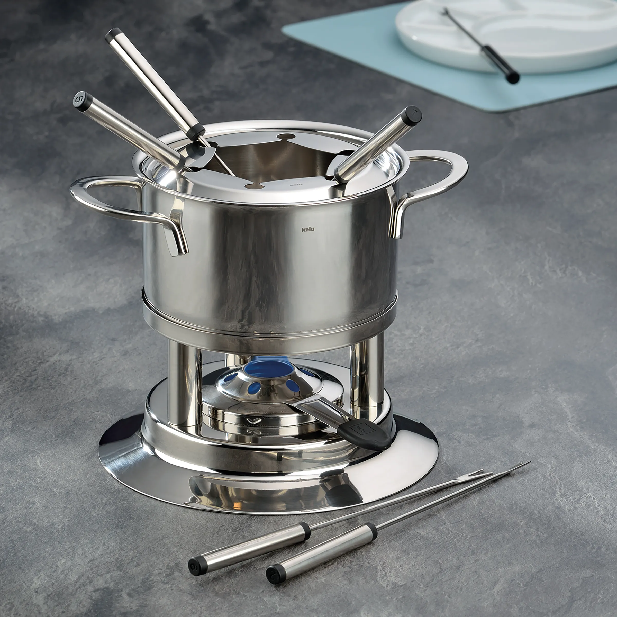 Kela Edelstahl-Fondue-Set mit Brenner, Topf, Deckel und sechs Gabeln auf grauer Arbeitsplatte