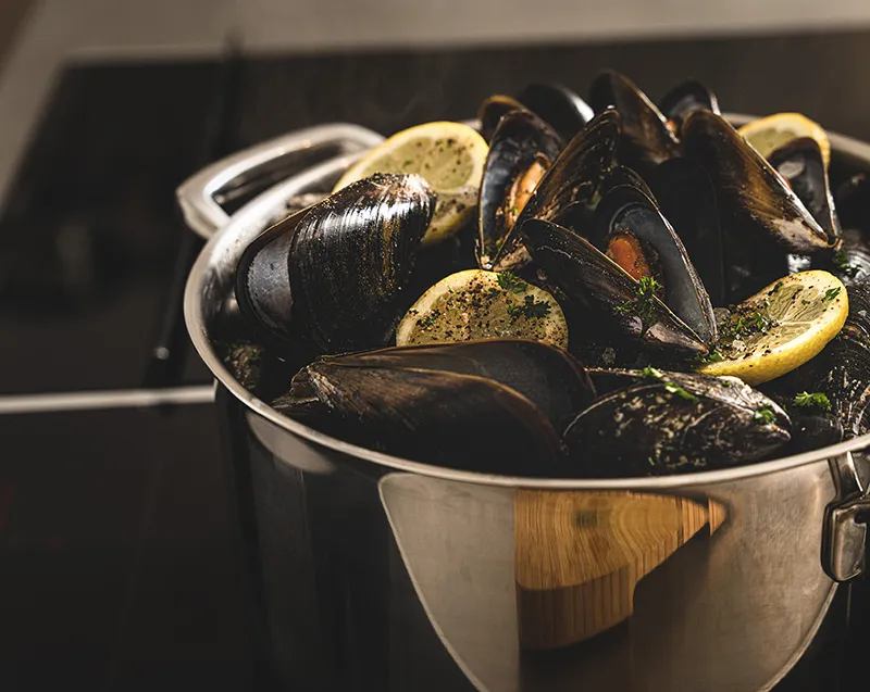 Miesmuscheln kochen im Topf mit Zitrone und Kräutern Schwarze Miesmuscheln mit Zitronenscheiben und Kräutern in einem Edelstahltopf auf dem Herd