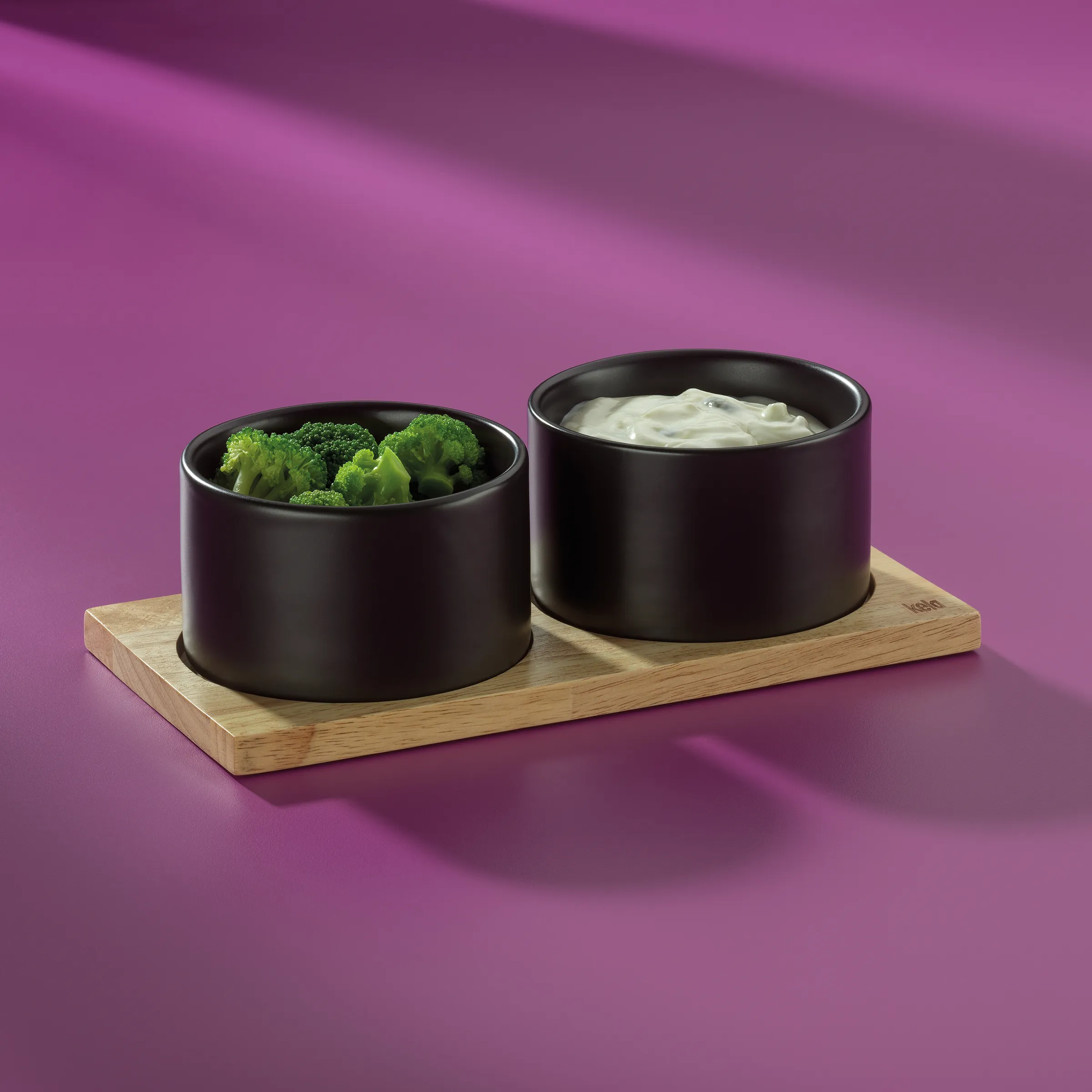 Zwei schwarze Keramikschüsseln auf Holzbrett mit Broccoli und Dip vor violettem Hintergrund