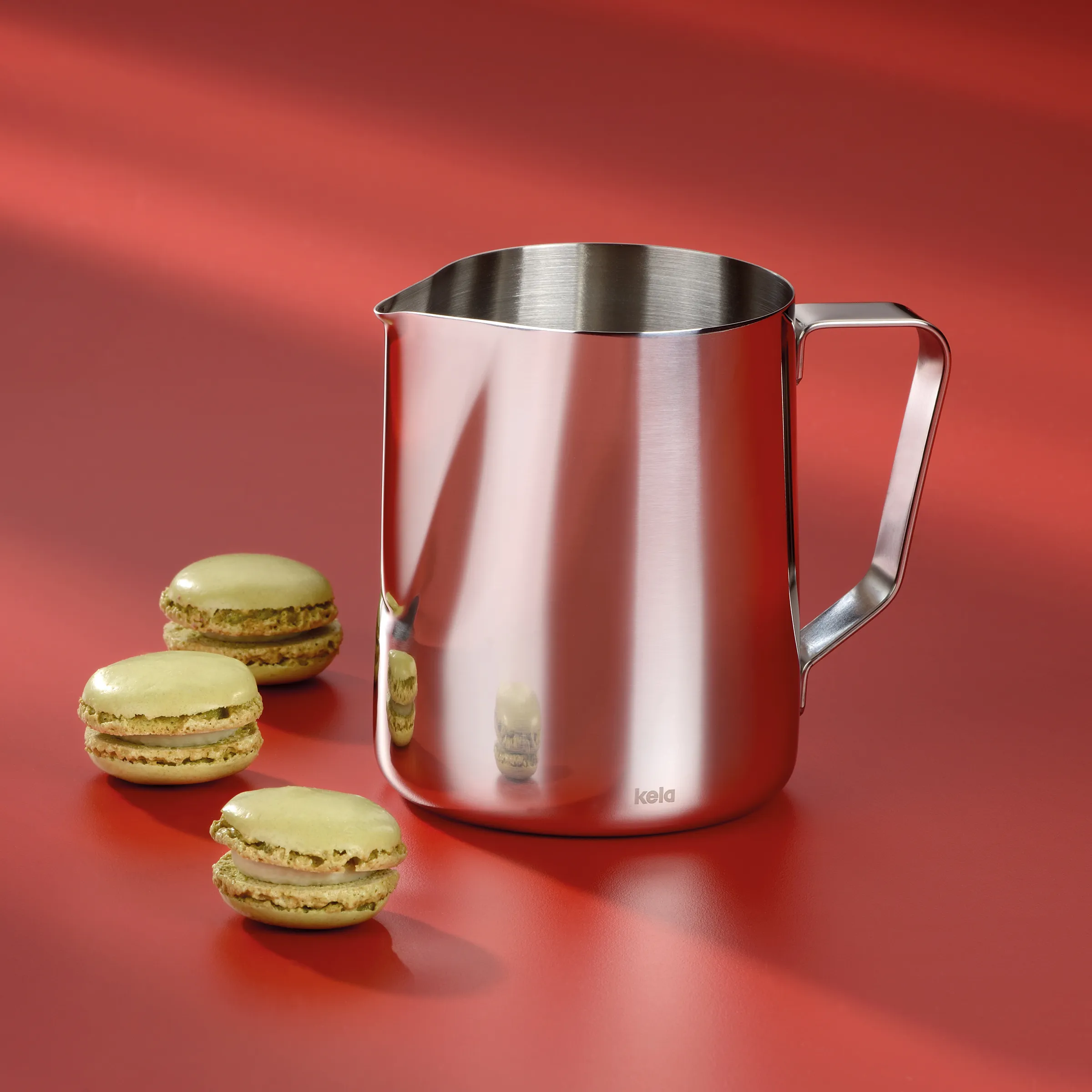 Edelstahl-Milchkännchen von kela mit drei hellen Macarons auf rotem Hintergrund