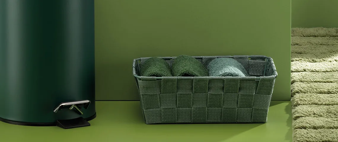 Grüner Aufbewahrungskorb mit Handtüchern und Zubehör Grüner Korb mit grünen Handtüchern, schwarze Schaufel und gerollte Handtücher auf grünem Hintergrund