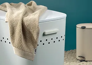 Wäschekorb Weiß - Moderne Wäschebox mit Deckel Weißer Wäschekorb mit Henkel und gelber Decke auf türkisfarbenem Hintergrund neben beiger Kerze