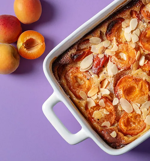 Aprikosenkuchen Rezept mit Mandeln Aprikosenkuchen in weißer Auflaufform mit Mandeln und frischen Aprikosen auf violettem Hintergrund