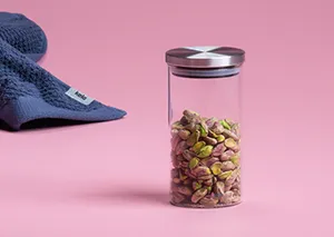 Transparenter Vorratsbehälter mit Metalldeckel gefüllt mit Müsli auf rosa Hintergrund neben blauem Tuch