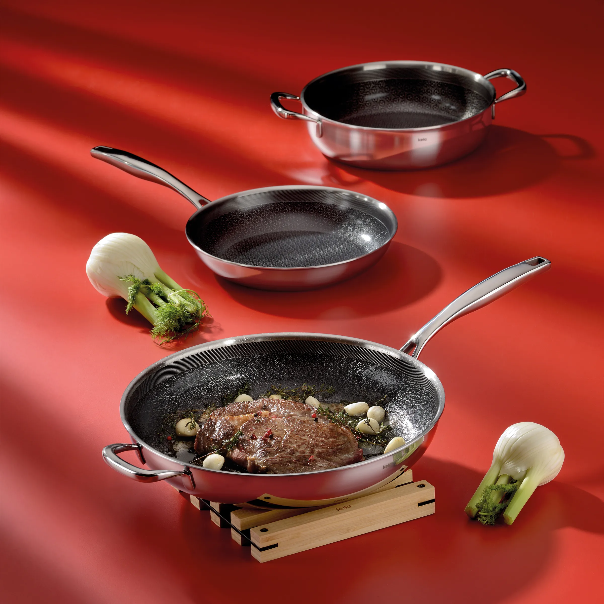 Kochset Pfannen und Töpfe - Hochwertiges Kochgeschirr Kochset mit Bratpfanne, Wokpfanne und Topf auf rotem Untergrund, daneben frisches Gemüse und Fleisch