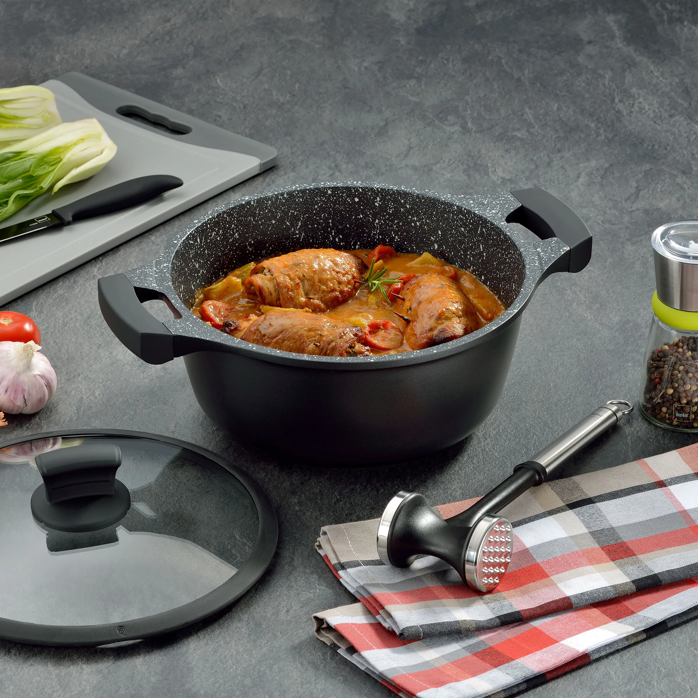 Schwarzer Kochtopf mit Deckel auf grauer Oberfläche, gefüllt mit gebratenen Hähnchenschenkeln in Tomatensauce und Kräutern.