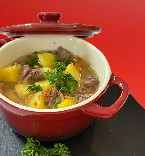 Rindergulasch mit Kartoffeln im Gusseisen-Schmortopf - Eintopf Rezept Rindergulasch mit Kartoffeln und Petersilie in rotem Gusseisen-Schmortopf auf dunkler Schieferplatte