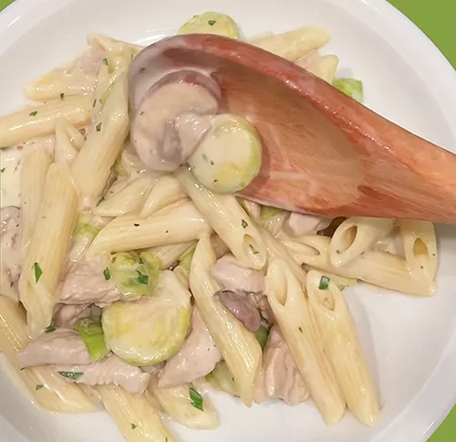 Penne Pasta mit Hähnchen, Zucchini und Zwiebeln auf weißem Teller mit Holzlöffel