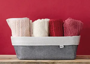 Grauer Wäschekorb mit weißem Rand gefüllt mit zusammengelegten Handtüchern in Rosa-, Creme- und Bordeauxtönen vor roter Wand