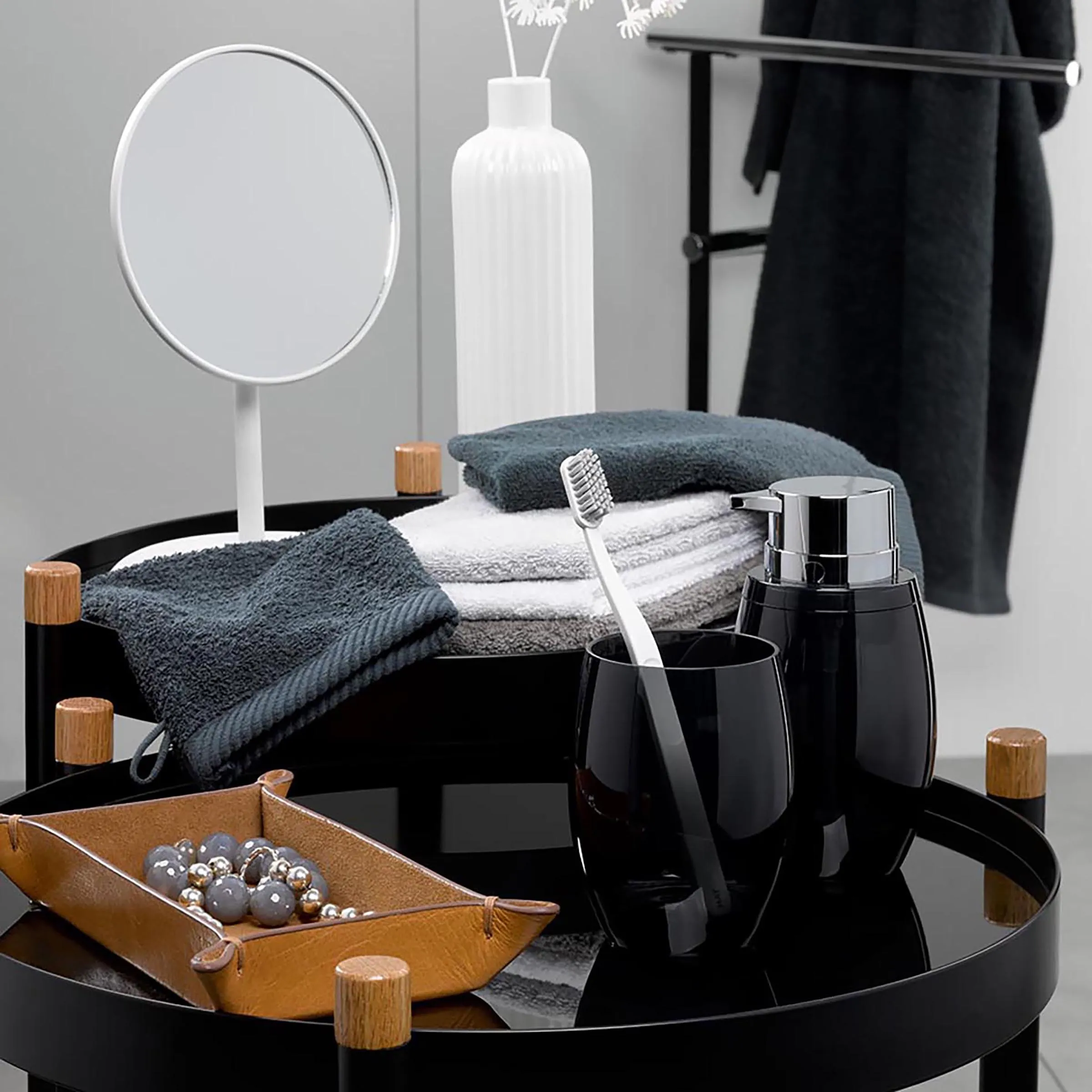 Badezimmer Accessoires Set - Moderne Badausstattung Modernes Badezimmer-Set mit schwarzem Tisch, weißem Spiegel, grauen Handtüchern, schwarzen Behältern und brauner Schale mit Dekokugeln