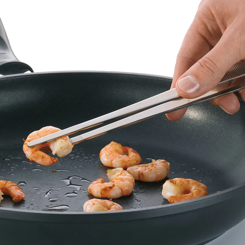 Garnelen Zange Pfanne Kochen Person mit Metallzange hält gebratene Garnele in schwarzer Pfanne mit weiteren Garnelen