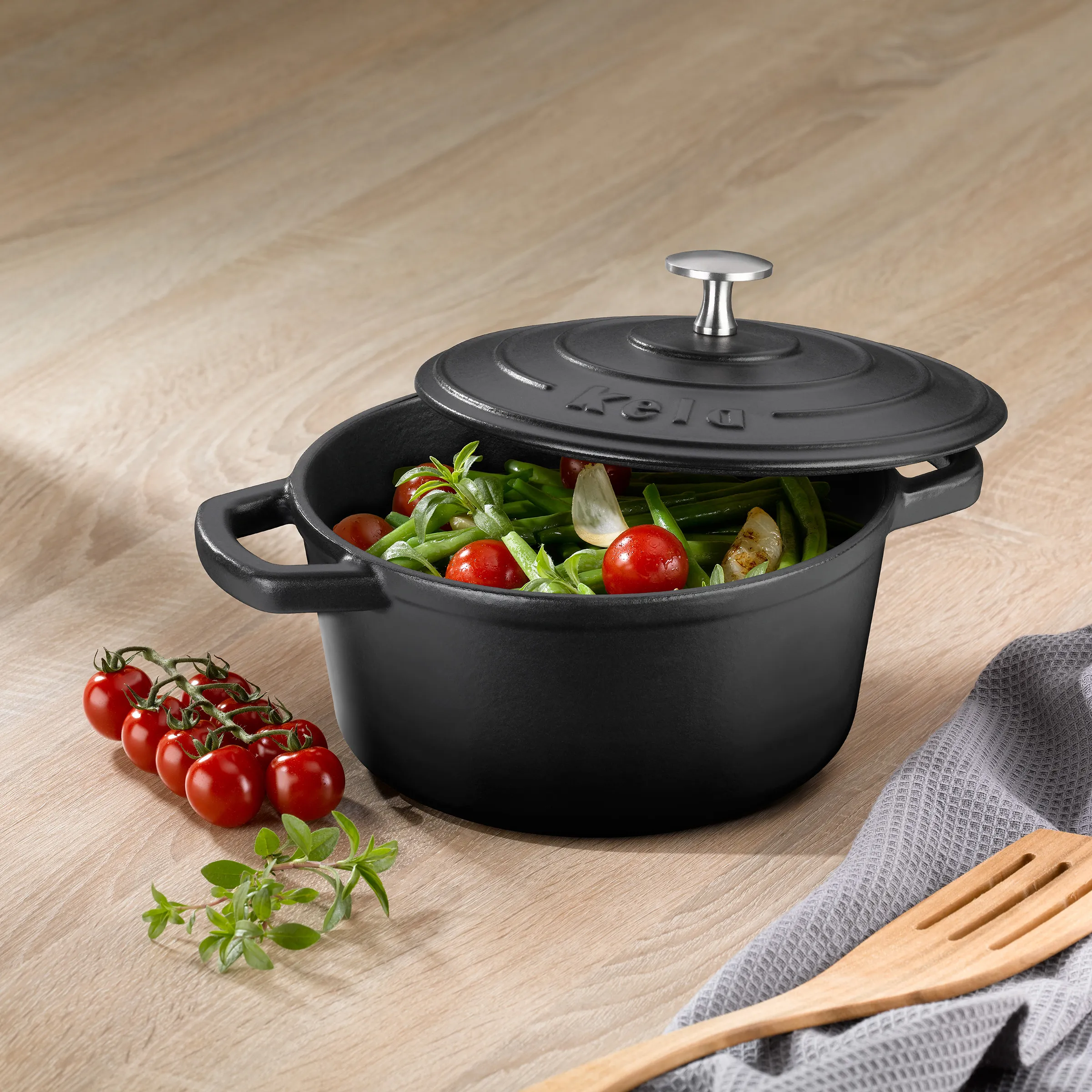 Gusseisen-Kochtopf mit Deckel und Gemüse - Hochwertiges Kochgeschirr Schwarzer Gusseisen-Kochtopf mit Deckel gefüllt mit frischem Gemüse auf Holztisch mit Tomaten und Kochutensilien