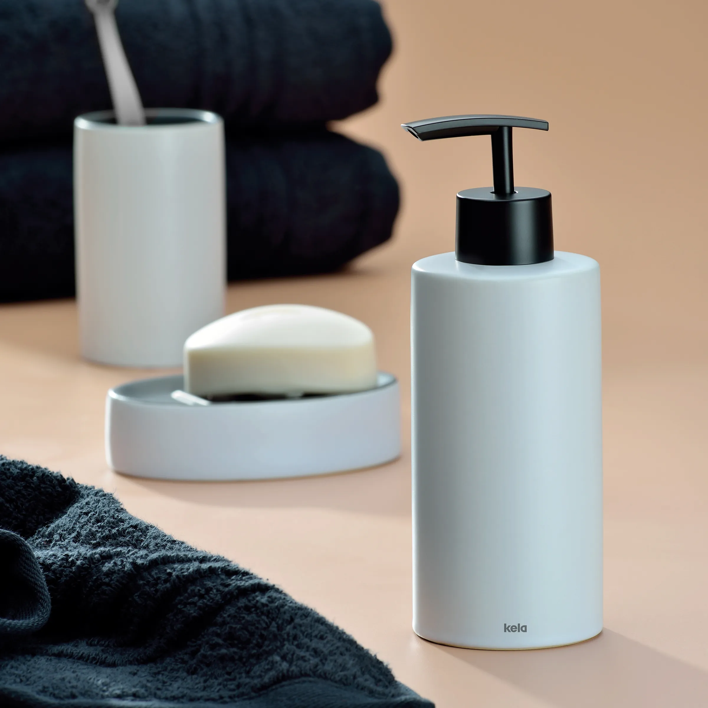 Badezimmer-Set mit weißem Seifenspender, Seifenschale, Zahnputzbecher und dunklem Handtuch auf beigem Hintergrund