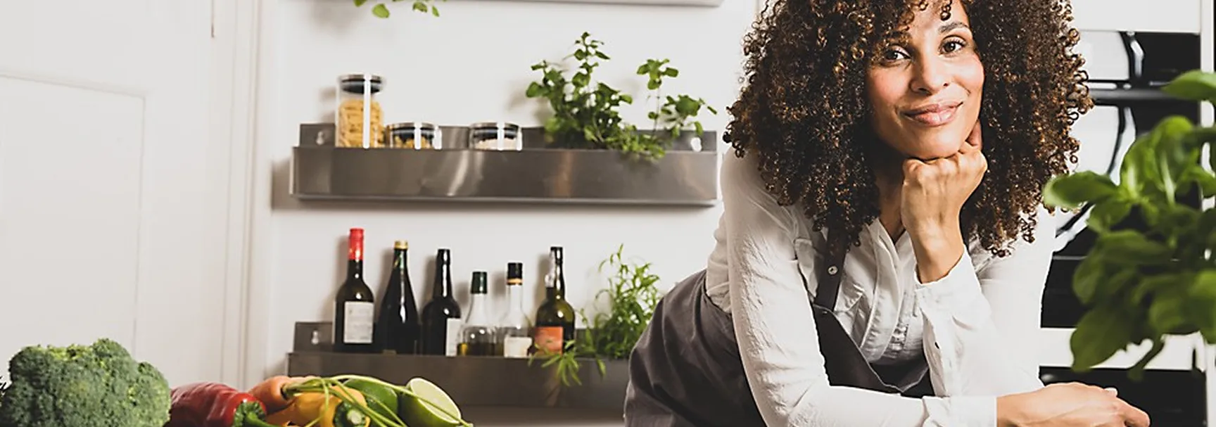 Modern kitchen design with fresh ingredients and herb plants Woman with curly hair smiling in modern kitchen with plants, fresh vegetables and wine bottles on shelf